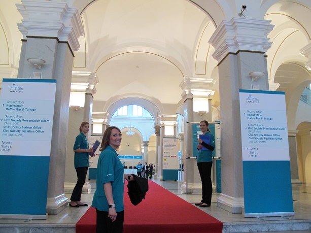 Event staff standing along a red carpet in the entrance hall of a conference venue, preparing to welcome guests to an international meeting.