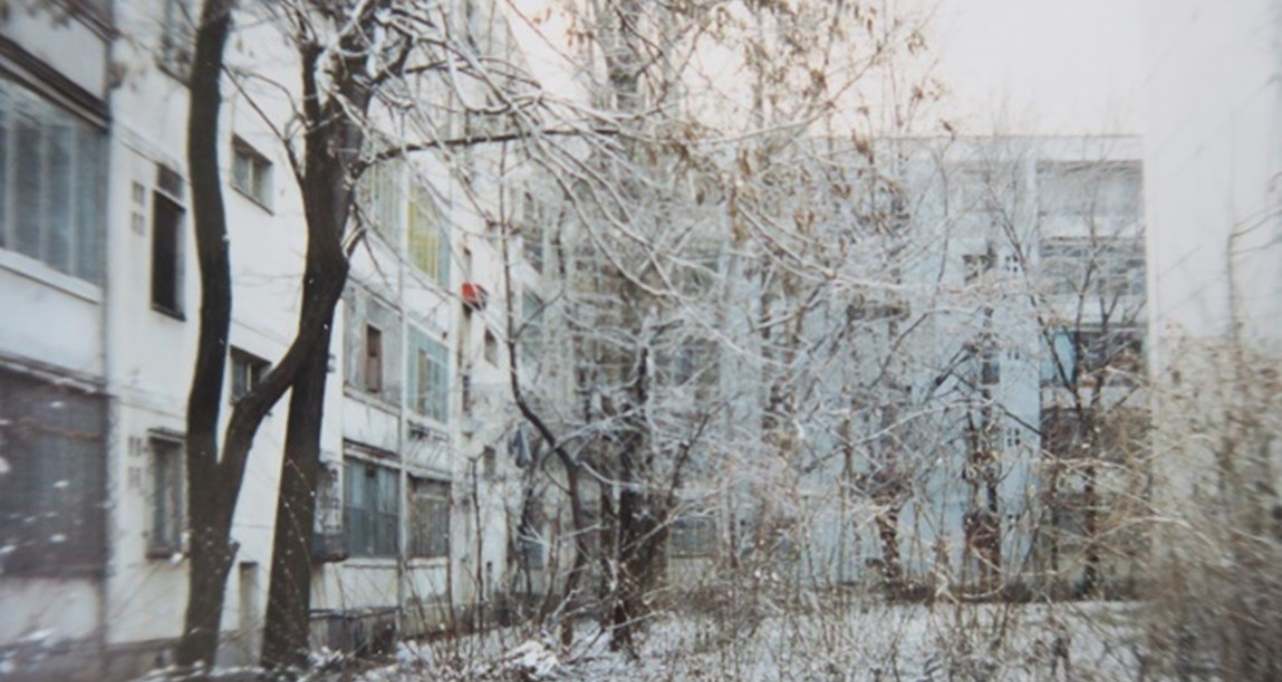 Winter view of a residential courtyard, with snow-covered trees and apartment buildings in the background.