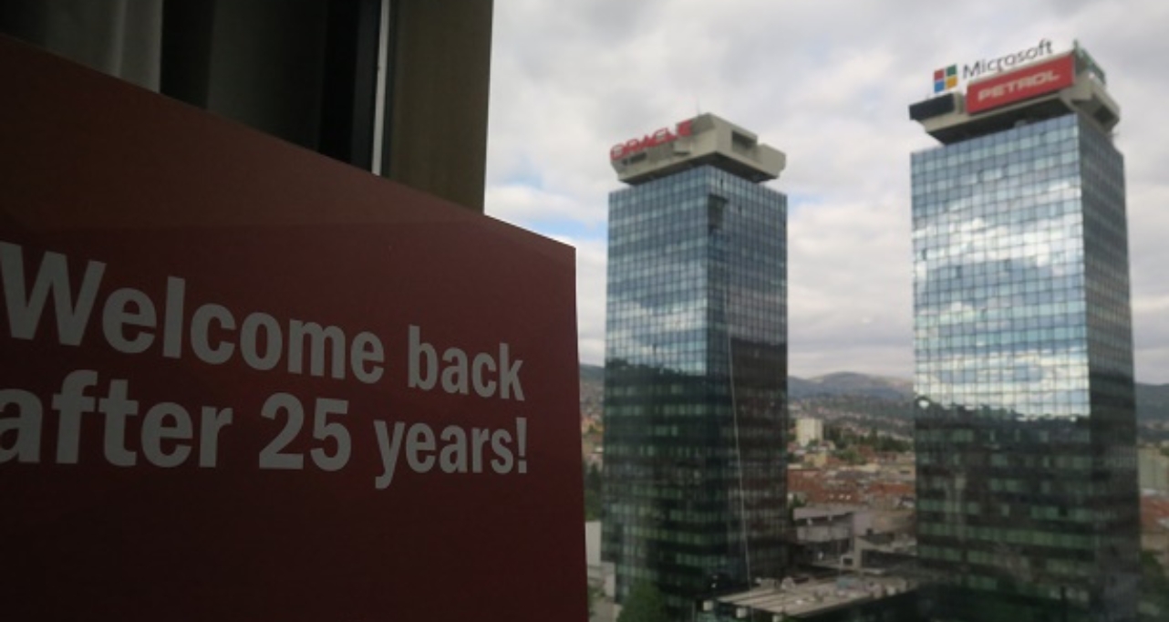 Welcome back after 25 years sign with a view of office towers through a window.