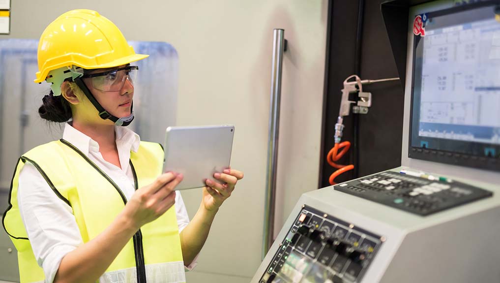 A female engineer wearing a yellow safety hat inspecting equipment using a tablet