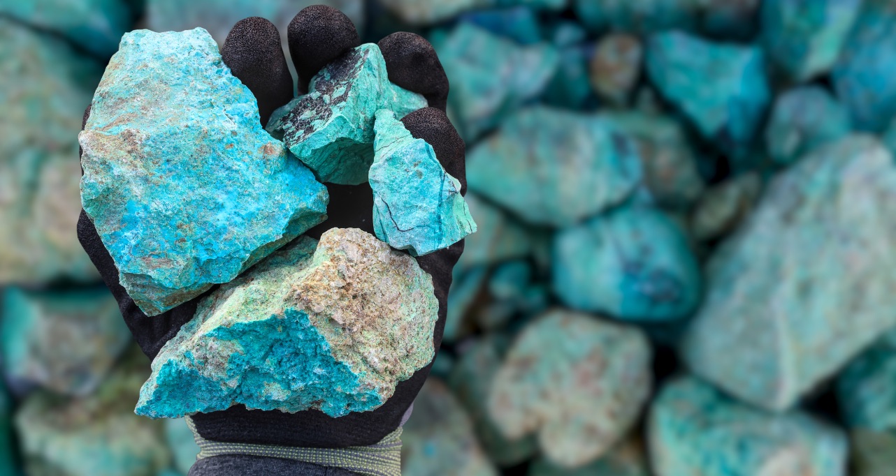 A black-gloved hand holds rough turquoise-blue mineral rocks above a pile of similar stones.